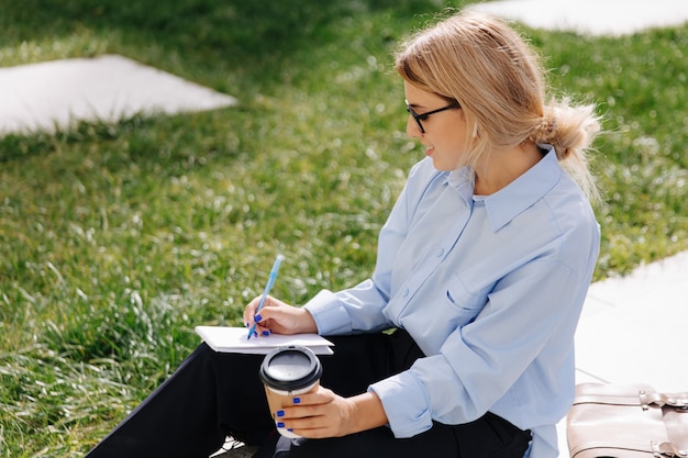 Young female hipster in blue shirt and black pants sitting outdoors and writing in notebook. Pretty woman in eyeglasses drinking coffee while studying on street.