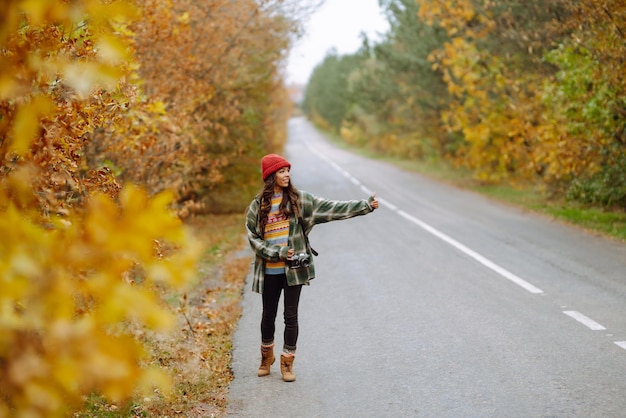 Young female hitchhiker by roadside among autumn forest during fall season Travel woman hitchhiking