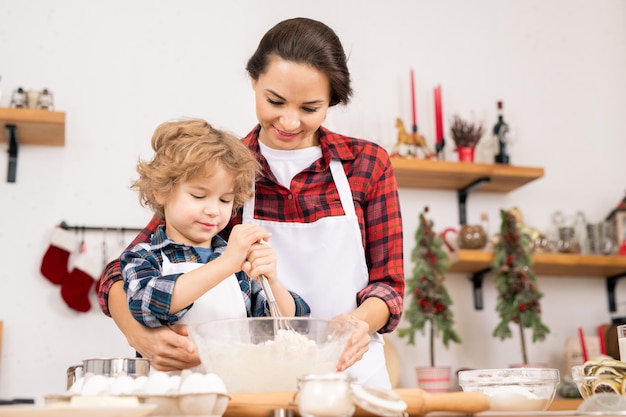 Young female holding bowl with flour and eggs while her son whisking them in process of preparing dough for homemade pastry