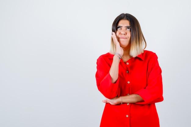 Young female holding hand on cheek in red oversized shirt and looking thoughtful , front view.