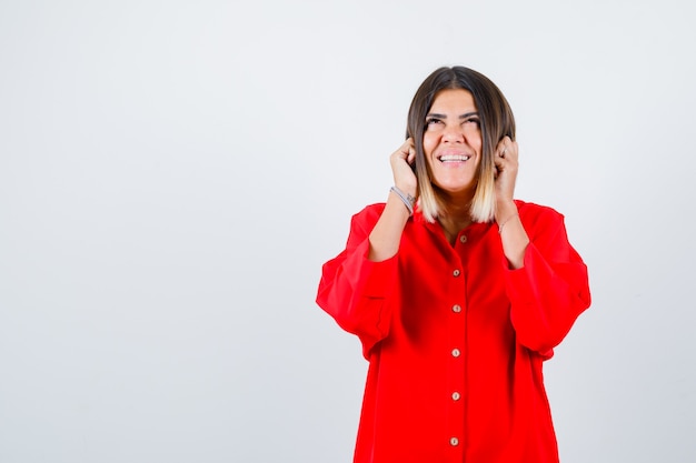 Giovane donna che si tiene per mano sulle guance in camicia rossa oversize e sembra allegra. vista frontale.