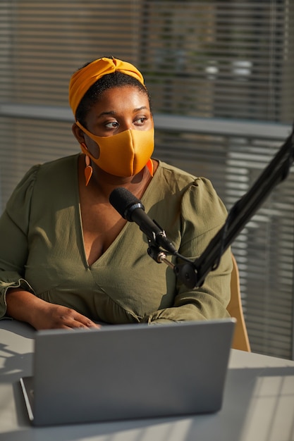 ラップトップを使用し、ラジオでの放送中にマイクで話す保護マスクの若い女性インタビュアー