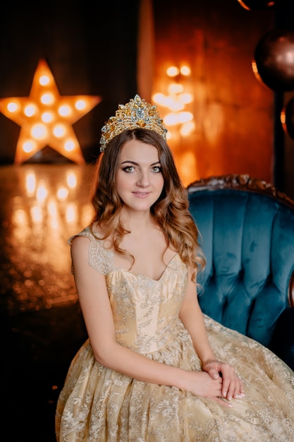 Young female is sitting in armchair in luxurious gold dress and looking into camera