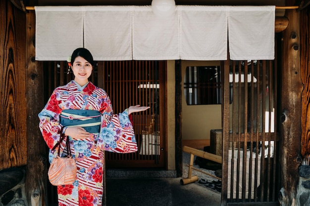 写真 カラフルな着物を着た若い日本人女性の妻が、伝統的な歴史ある温泉ホテルの木製のドアの前に立ち、歓迎の手のジェスチャーで笑顔のカメラを持っています。カーテンの下の花柄のドレスを着た女性