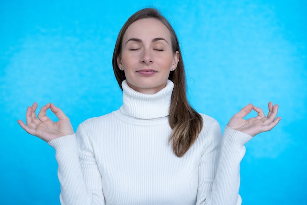 Young female keeping eyes closed while practicing yoga indoors, meditating, holding hands in mudra gesture