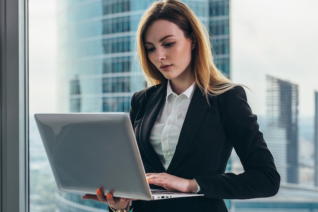 Young female lawyer working in her luxurious office holding a laptop standing against panoramic window with a view on business district.