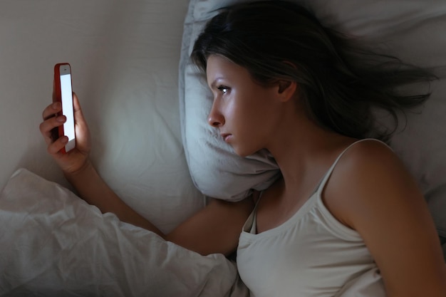 ベッドに横になり、夜に彼女のスマートフォンを見ている若い女性
