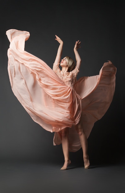 Young female in light chiffon dress standing with hands up dancing on black plain background.