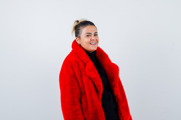 Foto giovane femmina che guarda l'obbiettivo in maglione, cappotto di pelliccia rosso e che sembra felice, vista frontale.