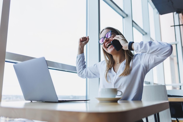 A young female manager in glasses works at a laptop and gesticulates emotionally with her hands while talking on the phone she is sitting at her desk in a white shirt