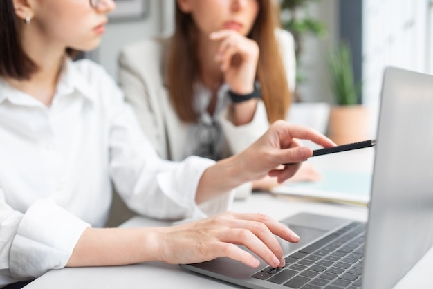 Young female manager showing something to colleague with pan pointing at laptop screen in office