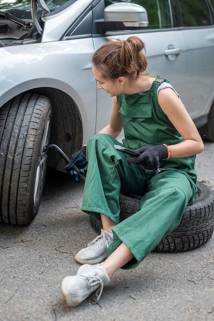 Young female mechanic in green uniform standing near a brokendown car and waiting help outdoor