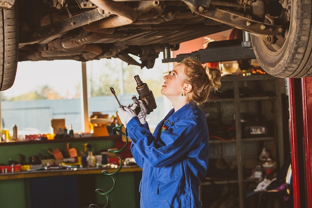 Young female mechanic with blonde hair in blue overall working under the car lifted on the hydraulic ramp fixing something