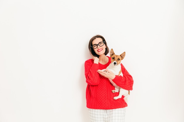 Young female model with glad expression, wears spectacles and red sweater, holds her favourite jack russell terrier dog