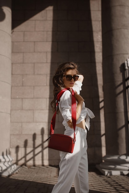 Photo young female model with perfect slim body in trendy sunglasses and white pants and shirt walking on the sidewalk of the european city