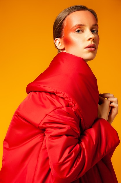 Young female modern wrapping in warm oversize red coat and looking at camera against yellow background