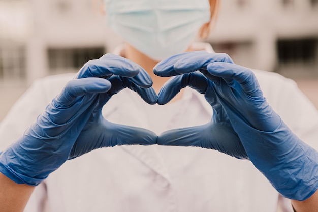 Young female nurse standing outside hospital and showing heart sign