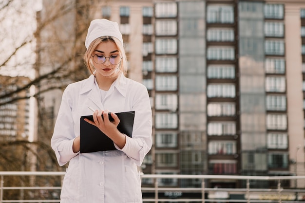 Young female nurse standing outside hospital and writing\
patient illness history in journal