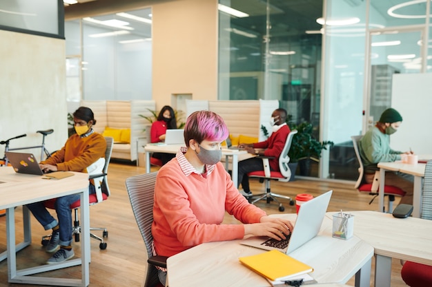 Young female office manager in casualwear and protective mask looking at laptop display while preparing presentation against her colleagues