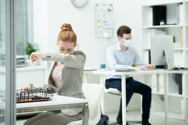 Young female office manager in suit and protective mask putting her face on elbow while going to sneeze by workplace against male colleague