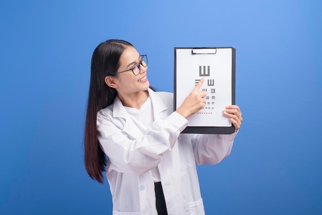 A young female ophthalmologist with glasses holding eye chart over blue background studio, healthcare concept