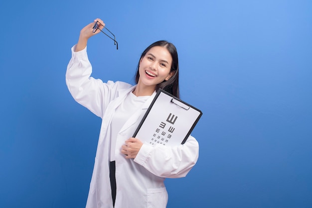 A young female ophthalmologist with glasses holding eye chart over blue wall