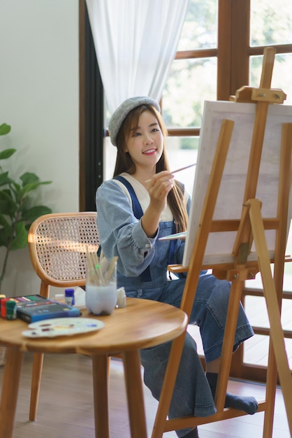 젊은 여성 화가는 컬러 팔레트를 들고 브러시를 사용하여 홈 스튜디오의 캔버스에 그림을 그립니다.
