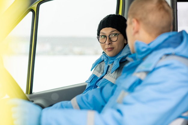 Young female paramedic in uniform and cap talking to driver of ambulance car while hurrying to save sick person