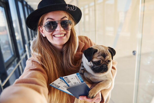 Young female passenger in warm clothes holding tickets and cute dog in hands in airport hall.