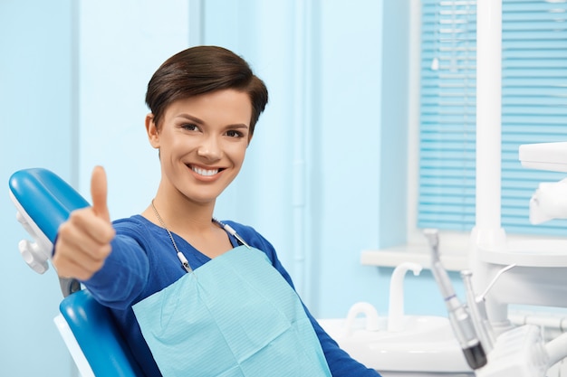 Ufficio dentista di visita del giovane paziente femminile. bella donna sorridente con i denti bianchi diritti sani che si siedono alla sedia dentale e che compongono i pollici. clinica dentale. stomatologia