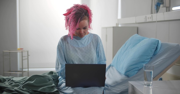 Young female patient working with laptop sitting on bed in hospital room