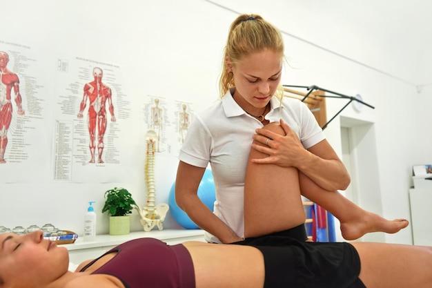 Young female physiotherapist exercising with her patient laying on massage table, flexing her knee muscles
