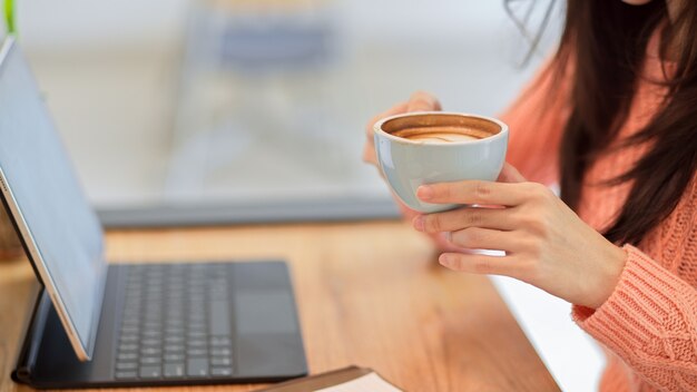 Молодая женщина в розовом свитере пьет, держа чашку горячего кофе, работая над планшетом в кафе
