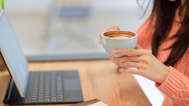 カフェでタブレットに取り組んでいる間、ホットコーヒーカップを保持して飲んでピンクのセーターを着た若い女性