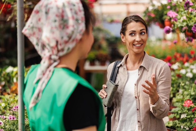 Young female plant nursery volunteer helps a young woman with flowers choosing and purchase.