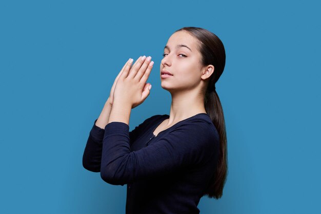 Photo young female pleading holding hands in prayer on blue background profile view