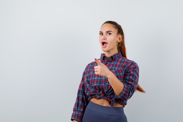 Young female pointing away in checkered shirt, pants and looking puzzled. front view.