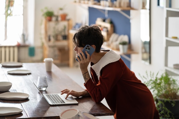 Foto giovane imprenditore femminile di ceramiche che utilizza il computer portatile che parla con il cliente tramite telefono controllando l'ordine online