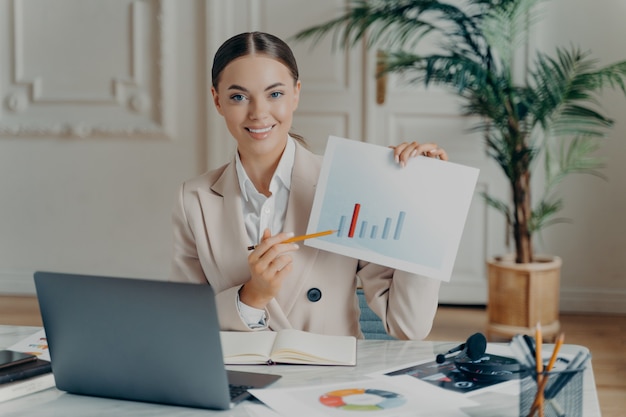 Foto giovane project manager o economista femminile con i capelli legati in coda di cavallo che punta al rapporto finanziario con statistiche in aumento e guardando la telecamera, seduta sul posto di lavoro con il laptop aperto in un ufficio accogliente