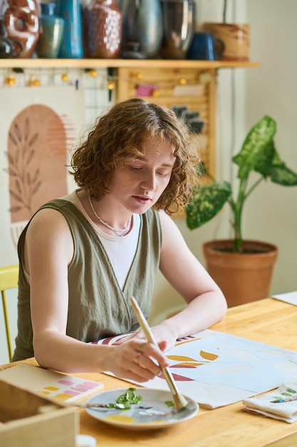 Young female putting paintbrush in plate with mixed gouache while painting