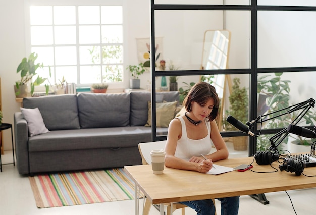 Young female radio host in casualwear making notes on paper by\
workplace