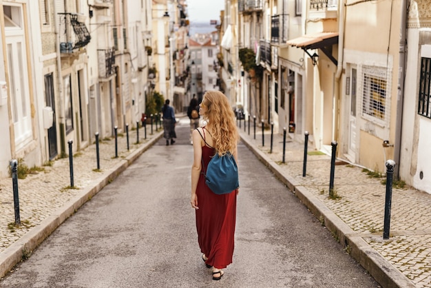 Young female in a red dress walking through a road surrounded by buildings under the sunlight