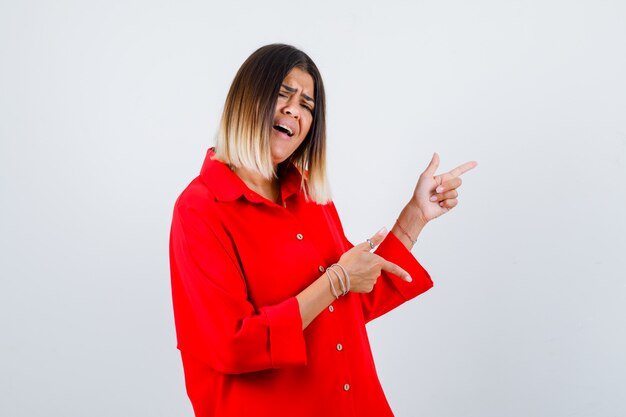 Young female in red oversized shirt pointing to the right side and looking energetic , front view.