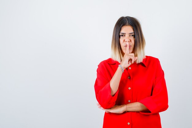 Young female in red oversized shirt showing silence gesture and looking confident , front view.