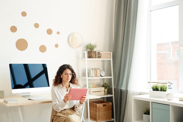 Young female remote worker sitting on chair in her room taking part in online meeting using her tablet computer