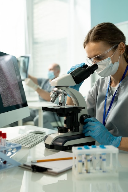 Young female scientist or clinician in uniform, protective mask, gloves and eyewear looking at chemical samples in microscope in laboratory