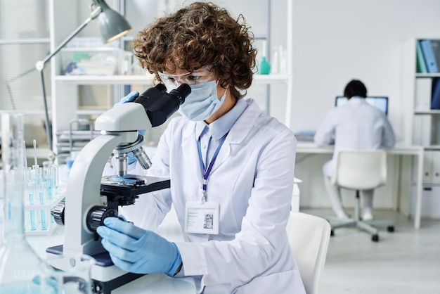 Young female scientist in protective mask and gloves looking in microscope