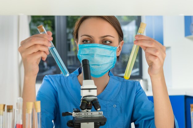 Young female scientist in uniform wearing a mask is holding test tubes with liquid and making some research in a laboratory. Healthcare and biotechnology concept