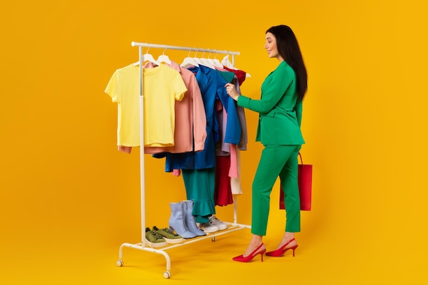 Young female shopaholic shopping and picking outfit choosing colorful clothes on rail standing over yellow background