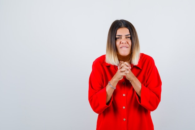 Young female showing clasped hands in pleading gesture in red oversized shirt and looking hopeful. front view.